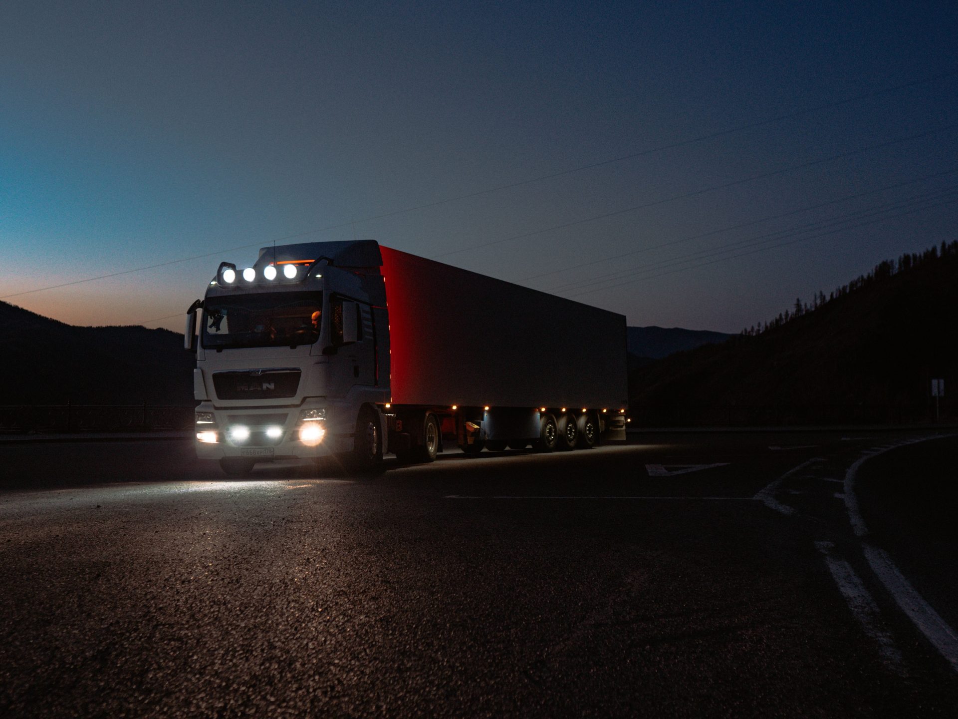 A heavy-duty truck navigates a quiet road at dusk, highlighting its illuminated path through remote mountains.
